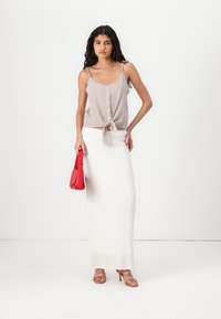 Top sin mangas de rayas en beige claro con un nudo frontal, combinado con una falda maxi blanca texturizada. Sostiene un pequeño bolso rojo.