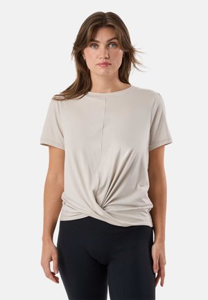 Woman with shoulder-length brown hair wearing a beige short-sleeve top with front knot detail and black leggings, facing forward.