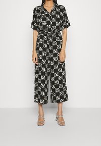Combinaison noire et blanche à motifs, manches courtes, taille ceinturée et pantalon à jambes larges. Portée avec des sandales à talons blocs et brides.