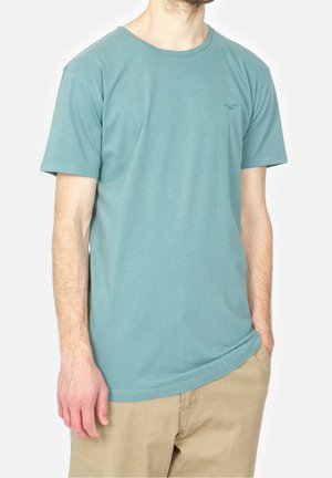 Mężczyzna w prostym turkusowym t-shircie z krótkim rękawem i małym logo na piersi, w połączeniu z beżowymi spodniami, ręce swobodnie opuszczone wzdłuż ciała.