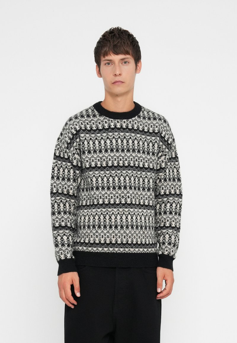 Μαύρο και άσπρο πλεκτό πουλόβερ με στρογγυλή λαιμόκοψη, μακριά μανίκια και ριπ σιρίτια και στρίφωμα. Παρουσιάζει περίπλοκα γεωμετρικά σχέδια.