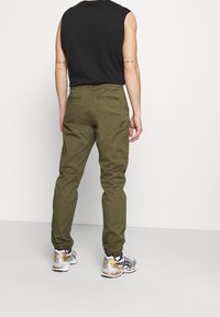 Olijfgroene chinos met een rechte pasvorm, twee achterzakken en een zachte textuur, gecombineerd met zilveren en gouden sport schoenen.