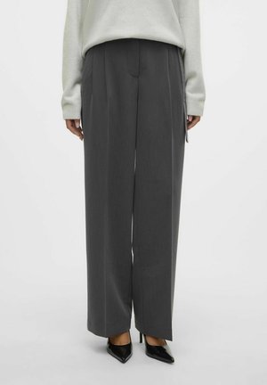 Pantalons larges gris avec une finition texturée, présentant des plis sur le devant, des poches latérales et une silhouette ajustée, associés à des talons noirs.