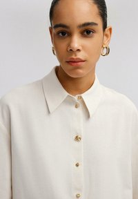 Camicia bianca con colletto classico, dotata di bottoni dorati e una texture di tessuto liscia. Il modello indossa orecchini a cerchio.