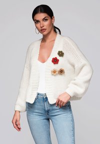 Vit stickad kofta med lös passform, prydd med färgglada tygblommor i röd, grön och beige på framsidan.
