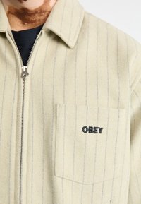 Jachetă striped de culoare bej deschis, confecționată dintr-un material moale. Dispune de un fermoar frontal, guler și un buzunar pe piept cu "OBEY" brodat cu negru.