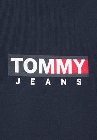 Tela de algodón azul marino con un logotipo rectangular que dice "TOMMY" en rojo y "JEANS" en blanco, embotellado sobre un fondo texturizado.