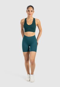 Een model in een donkergroene sportbeha en bijpassende short staat met de handen op de heupen tegen een neutrale achtergrond en kijkt zelfverzekerd naar de camera.