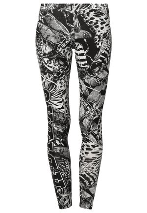 Persoon die een zwart ritsjack draagt, een wit lang T-shirt, zwart-wit patroonleggings en witte Adidas hoge sneakers.