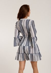 Weißes Kleid mit blauen geometrischen Mustern, peplum Saum und Glockenärmeln. Leichter Stoff mit strukturierter Oberfläche.