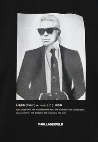 Fekete póló, amelyen egy napszemüveget viselő férfi látható öltönyben. Az alatta lévő szöveg így szól: "i-kon", és Karl Lagerfeldet jellemzi.