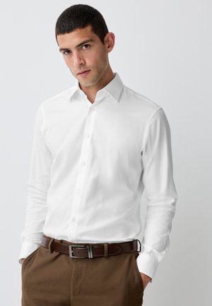 Uomo che indossa una camicia bianca con bottoni infilata in pantaloni marroni con una cintura di pelle marrone scuro, mani in tasca, su sfondo uniforme.
