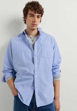Jonge man met krullend bruin haar die een lichtblauw gestreept overhemd draagt over een grijs shirt met lange mouwen en een blauwe spijkerbroek.