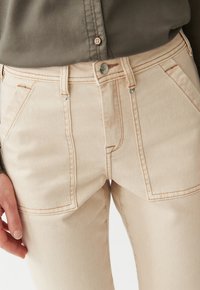 Light beige jeans featuring a fitted design, prominent front pockets, and contrasting stitching. The fabric has a smooth texture with a slight stretch.