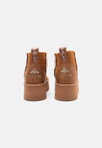 Brązowe zamszowe botki na platformie z ociepliną z baranka, oznaczone logo "REPLAY" na pięcie oraz tkaninowym uchwycie, widok z tyłu na białym tle.