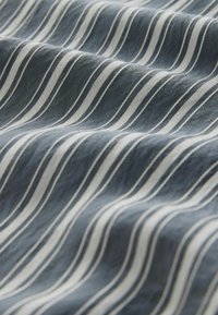 Modra tkanina z belimi valovitimi črtami, ki imajo mehko teksturo. Črte so enakomerno razporejene, kar ustvarja ritemski vzorec po celotni površini.