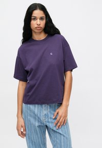 Violeta kokvilnas t-krekls ar brīvu piegriezumu, īsām piedurknēm un nelielu logo akcentu kreisajā krūtī; apvienots ar gaiši zilām svītrainām džinsiem.