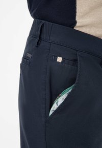 Navy blue pants made of textured fabric, featuring a zippered pocket with a green zipper and patterned interior, accented with two rear pockets.