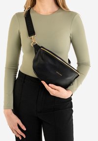 Czarna skórzana torebka na ramię o zakrzywionym kształcie, ze złotym suwakiem i drukowanym paskiem. Torebka noszona jest na oliwkowej zielonej koszuli.