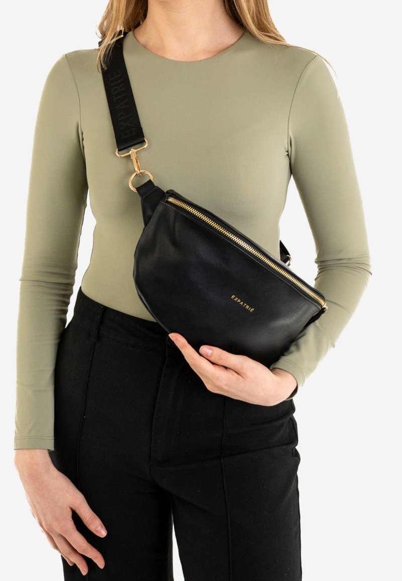 Czarna skórzana torebka na ramię o zakrzywionym kształcie, ze złotym suwakiem i drukowanym paskiem. Torebka noszona jest na oliwkowej zielonej koszuli.