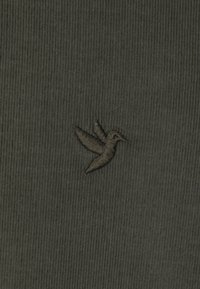 Dunkelgrüner Stoff mit einer vertikalen Rippstruktur zeigt ein kleines schwarzes gesticktes Kolibri-Logo auf der unteren linken Seite.