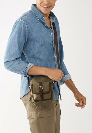 Sac à bandoulière kaki avec une surface texturée, deux poches avant et des accents de sangle brune. Porté sur une chemise en denim et un pantalon cargo.
