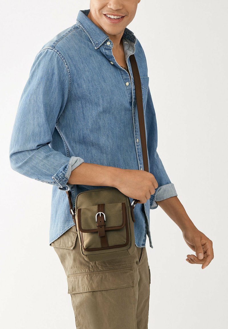 Sac à bandoulière kaki avec une surface texturée, deux poches avant et des accents de sangle brune. Porté sur une chemise en denim et un pantalon cargo.