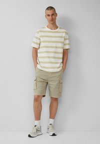 Weiß gestreiftes T-Shirt mit beige- und olivfarbenen Streifen, kombiniert mit beigen Cargo-Shorts mit seitlichen Taschen und hellen Sneakers mit Akzenten.