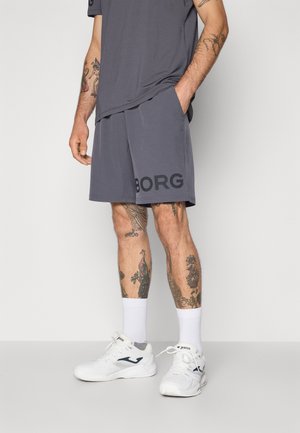 Shorts de sport gris avec une coupe ample, présentant un grand logo "BORG" sur la jambe gauche, associés à des chaussures de sport blanches et des chaussettes.