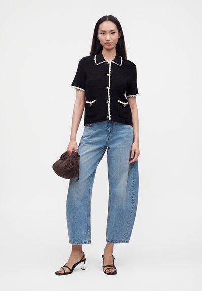 Femme portant un haut noir à manches courtes boutonné avec un bord blanc, jean bleu ample, sandales noires à talons, tenant un sac à main marron texturé.