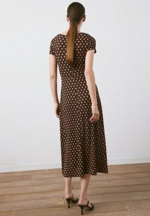 Femme aux longs cheveux bruns, portant une robe midi marron à pois clairs, des sandales à talons hauts noires, debout sur un sol en bois contre un mur blanc.