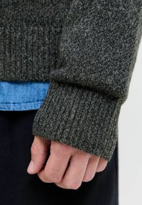 Manche de pull en maille gris foncé avec des poignets côtelés. Porté sur une chemise en denim bleu clair, mettant en valeur un tissu texturé.