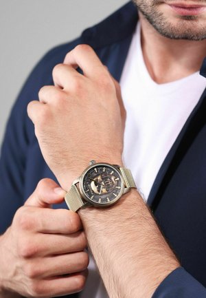 Montre en acier inoxydable avec un cadran noir et des accents dorés, dotée d'un bracelet en maille et d'un mécanisme interne visible, portée au poignet.