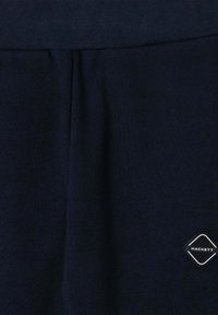 Tmavomodrá tkanina so stredovým švom a malou diamantovou nášivkou loga Hackett v bielej a čiernej farbe v pravom dolnom rohu.