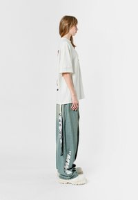 Camiseta de gran tamaño en gris claro con mangas enrolladas. Pantalones verdes holgados con detalles gráficos. Zapatos de plataforma gruesa en blanco. Vista lateral.