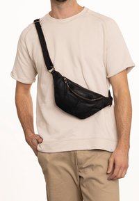 Mężczyzna w beżowej koszuli z krótkim rękawem i spodniach, z czarną skórzaną torbą na ramię, która ma zamek błyskawiczny z przodu oraz regulowany pasek.