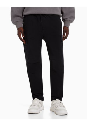 Mann trägt schwarze Cargohose mit seitlichen Taschen, grauen Pullover und weiße Turnschuhe, steht vor weißem Hintergrund.