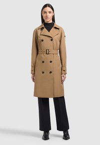 Donna che indossa un trench doppiopetto color sabbia con cintura, dolcevita nero, pantaloni neri a gamba larga e stivaletti neri, in piedi.