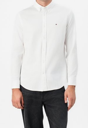 Camisa blanca de manga larga con botones y pequeño logo de Tommy Hilfiger en el pecho, llevada con pantalones gris oscuro.
