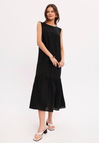 Czarna sukienka bez rękawów z warstwowym dołem, wykonana z lekkiego materiału. Charakteryzuje się luźnym krojem i subtelnym drapowaniem. Noszona w połączeniu z sandałami.