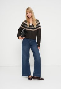 Cardigan maro închis cu un model Fair Isle la guler, purtat cu blugi din denim albastru cu croială largă și pantofi negri lejeri. Ține în mână o geantă de mână neagră.