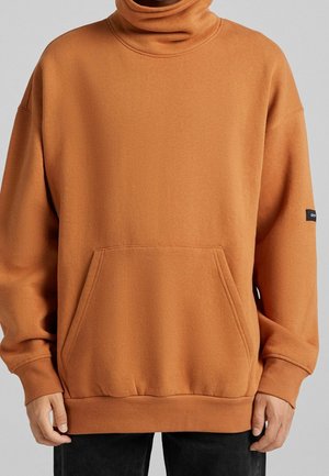 Maglione a collo alto color ruggine realizzato in morbido tessuto, con tasca frontale, polsini e orlo a costine, e un'etichetta sulla manica.