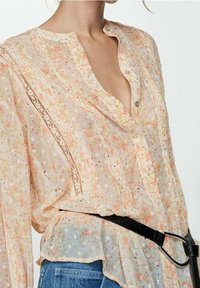 Blouse légère à fleurs en pêche doux avec des accents de dentelle, boutons sur le devant et manches fluides. Associée à une ceinture noire sur un jean en denim.