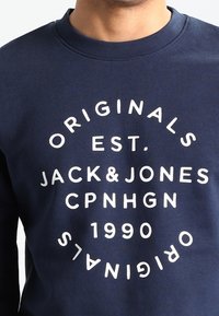 Královská modrá mikina s kulatým výstřihem, která obsahuje bílou grafiku s textem: "ORIGINALS EST. JACK&JONES CPNHGN 1990." Měkký materiál.