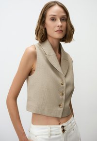 Beige verkürzter Weste aus strukturierter Stoff, mit Reverskragen und drei braunen Knöpfen. Kombiniert mit weißen, hochtaillierten Hosen.