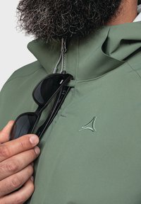 Giacca impermeabile verde con colletto alto, chiusura con zip e ricamo del logo discreto. Occhiali da sole neri tenuti in mano, che mostrano un design elegante.
