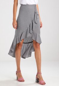 Schwarz-weißer Karorock mit asymmetrischem Saum und Rüschendetail, kombiniert mit rosa Absatzsandalen. Glatte Stoffstruktur.