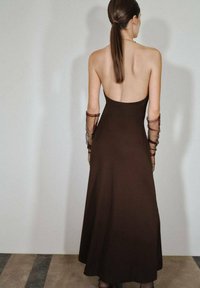 Brązowa, bezrękawna suknia z niskim tyłem, płynąca tkanina oraz przezroczyste, marszczone długie rękawy. Gładka tekstura i luźny krój.