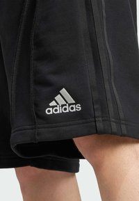 Crne atletske šortse sa tri bijele pruge uz strane, s bijelim Adidas logom na rubu. Napravljene su od glatkog, fleksibilnog materijala.