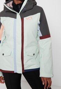 Lekka miętowo-zielona kurtka z szarymi, różowymi i bordowymi akcentami. Posiada kaptur, kieszenie na zamek oraz geometryczny wzór z blokami kolorów.